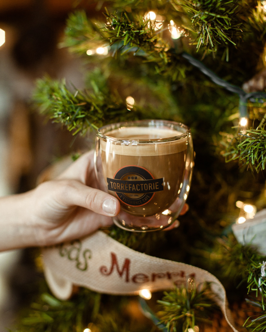 Verre à double paroi Torréfactorie tenu à la main devant un sapin, rempli d'un café lacté